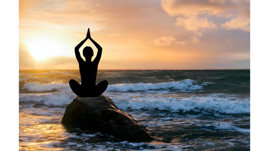 Woman practicing yoga on a rock by the beach during sunrise.